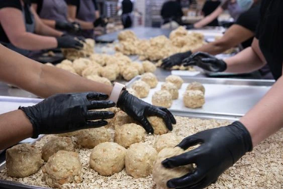 Photo of a cookie production scene with gloved hands shaping dough balls on trays