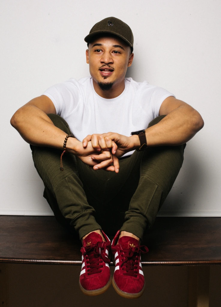 Sebastian Moy seated against a plain background wearing a white shirt, cap, and red sneakers